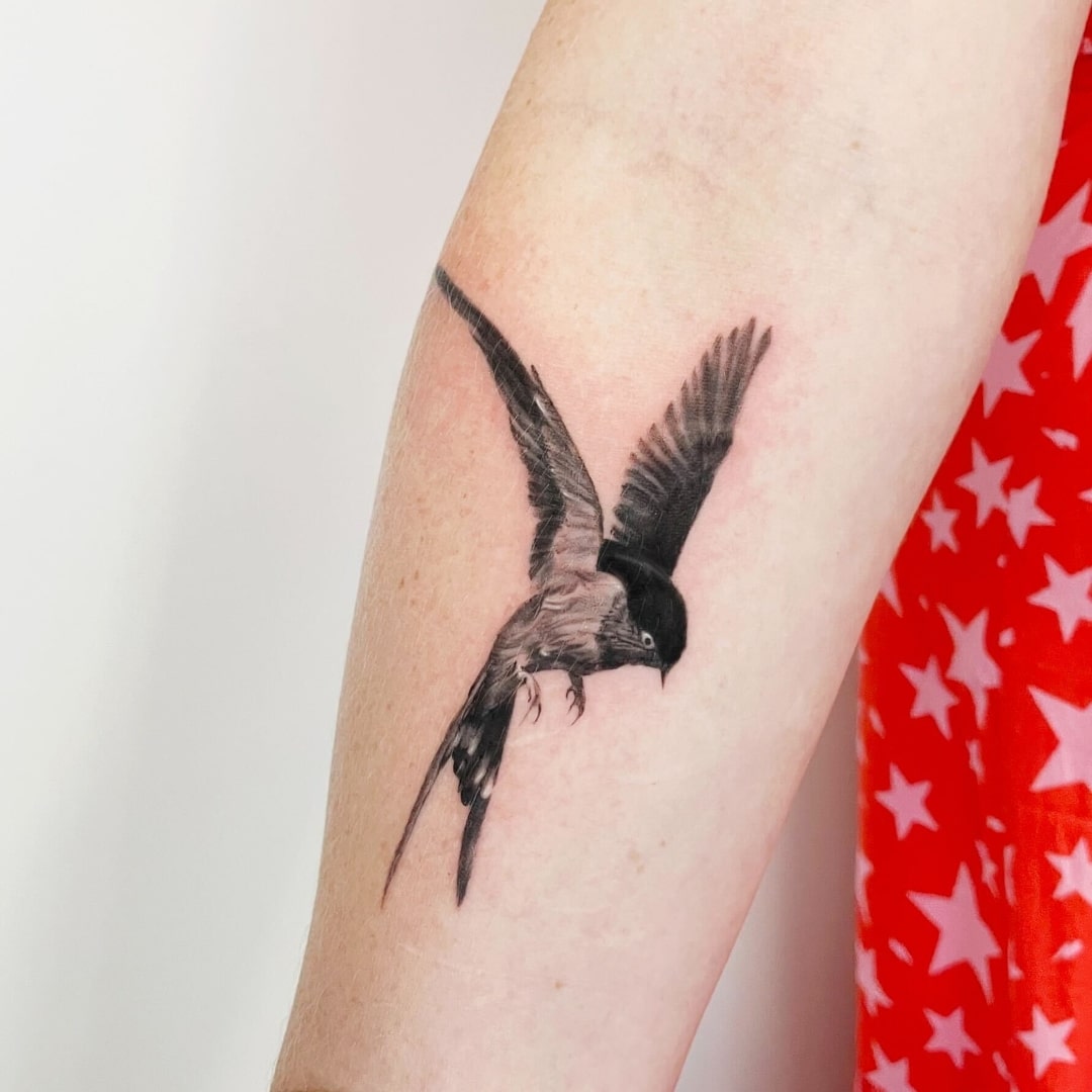 Close-up of a black and grey tattoo of a bird in flight on a forearm.