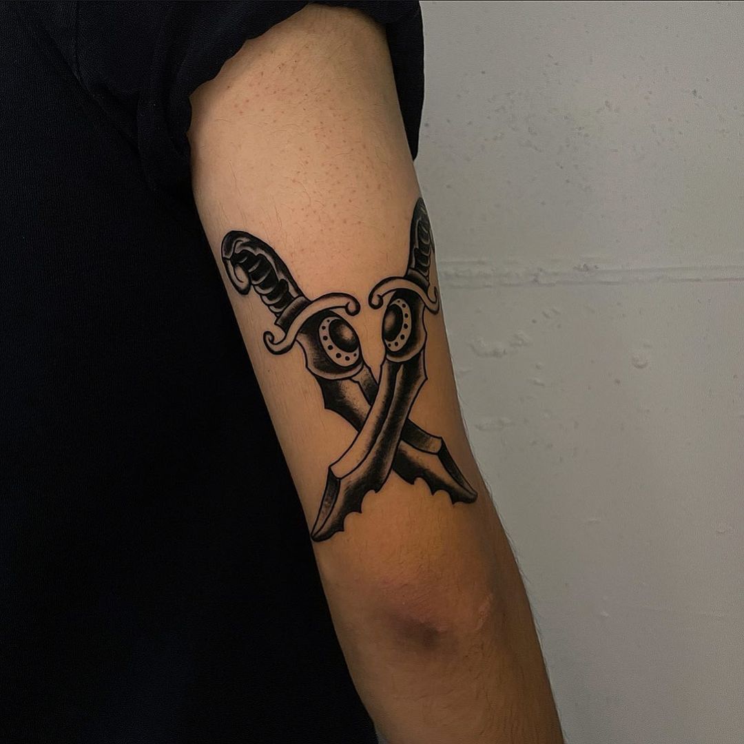 Black and grey tattoo of crossed daggers on arm.